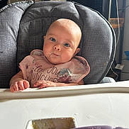 Olive is registered to the contest to win money with this photo: baby, high_chair, bib, food, spoon, bowl, indoor, child, blue_eyes, face, feeding, table, curious, messy, person, kitchen, furniture, clothing, infant, expression
