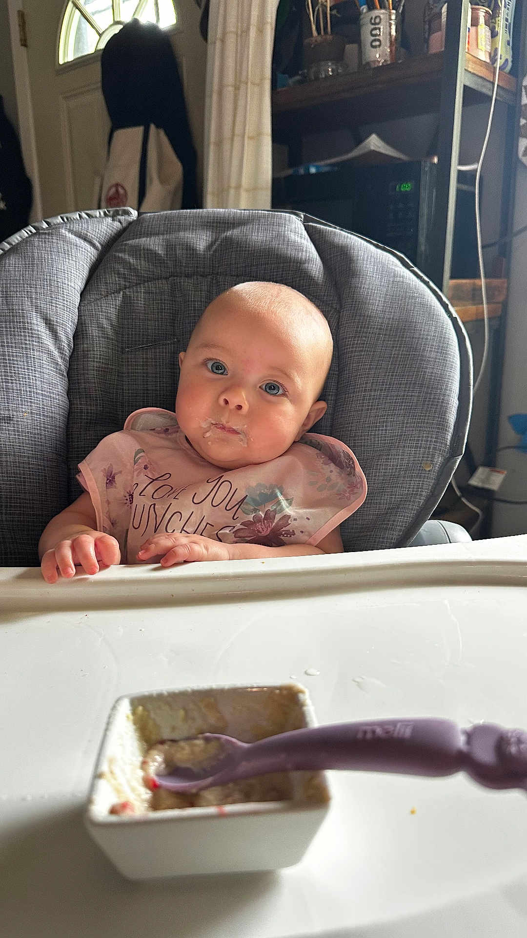 Olive is registered to the contest to win money with this photo: baby, high_chair, bib, food, spoon, bowl, indoor, child, blue_eyes, face, feeding, table, curious, messy, person, kitchen, furniture, clothing, infant, expression