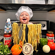 Noah is registered to the contest to win money with this photo: baby, child, infant, smiling, chef_hat, pasta, noodles, cooking, kitchen, food, spaghetti, vegetables, tomato, bell_pepper, onion, pasta_sauce, plush_toy, stovetop, portrait, six_months