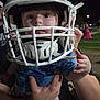 Andrew is registered to the contest to win money with this photo: child, baby, football_helmet, helmet, sports_equipment, night, outdoor, grass, person, hand, blue_clothing, adult_hand, face, eyes, headgear, protection, field, sports, cute, holding