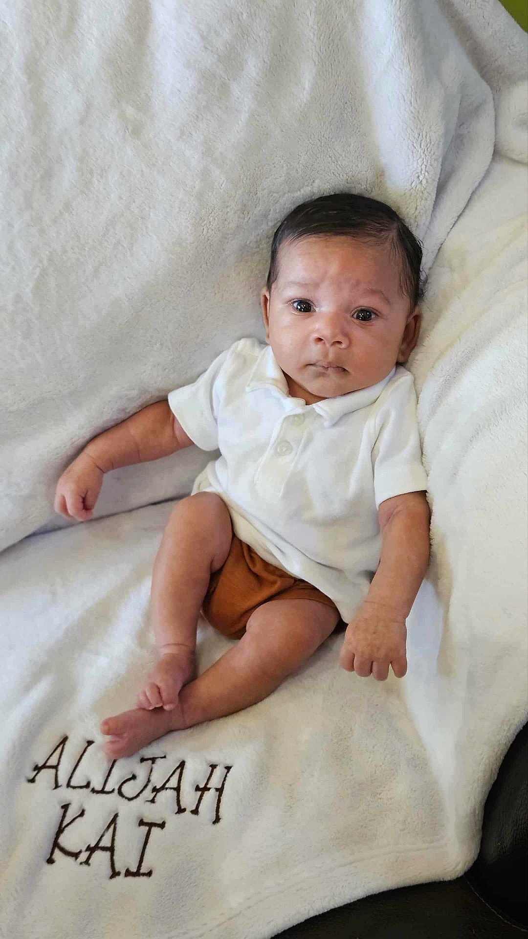 Alijah is registered to the contest to win money with this photo: baby, infant, child, blanket, white, polo_shirt, brown_shorts, face, eyes, expression, sitting, cozy, plush, soft, embroidery, name, cute, small, indoors, portrait