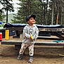 child, boy, hoodie, cap, outdoor, forest, picnic_table, dirt, shoes, pants, yellow_object, trees, nature, casual_clothing, daylight, smiling, standing, playful, recreation, park