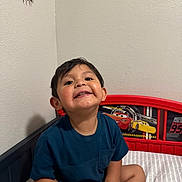 Emmanuel joined the competition — help win amazing prizes! child, smiling, bed, bedroom, tshirt, shorts, happy, face, person, indoor, wall, headboard, cartoon, toy_theme, young_child, sitting, casual_clothing, cute, home, playful