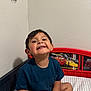 child, smiling, bed, bedroom, tshirt, shorts, happy, face, person, indoor, wall, headboard, cartoon, toy_theme, young_child, sitting, casual_clothing, cute, home, playful