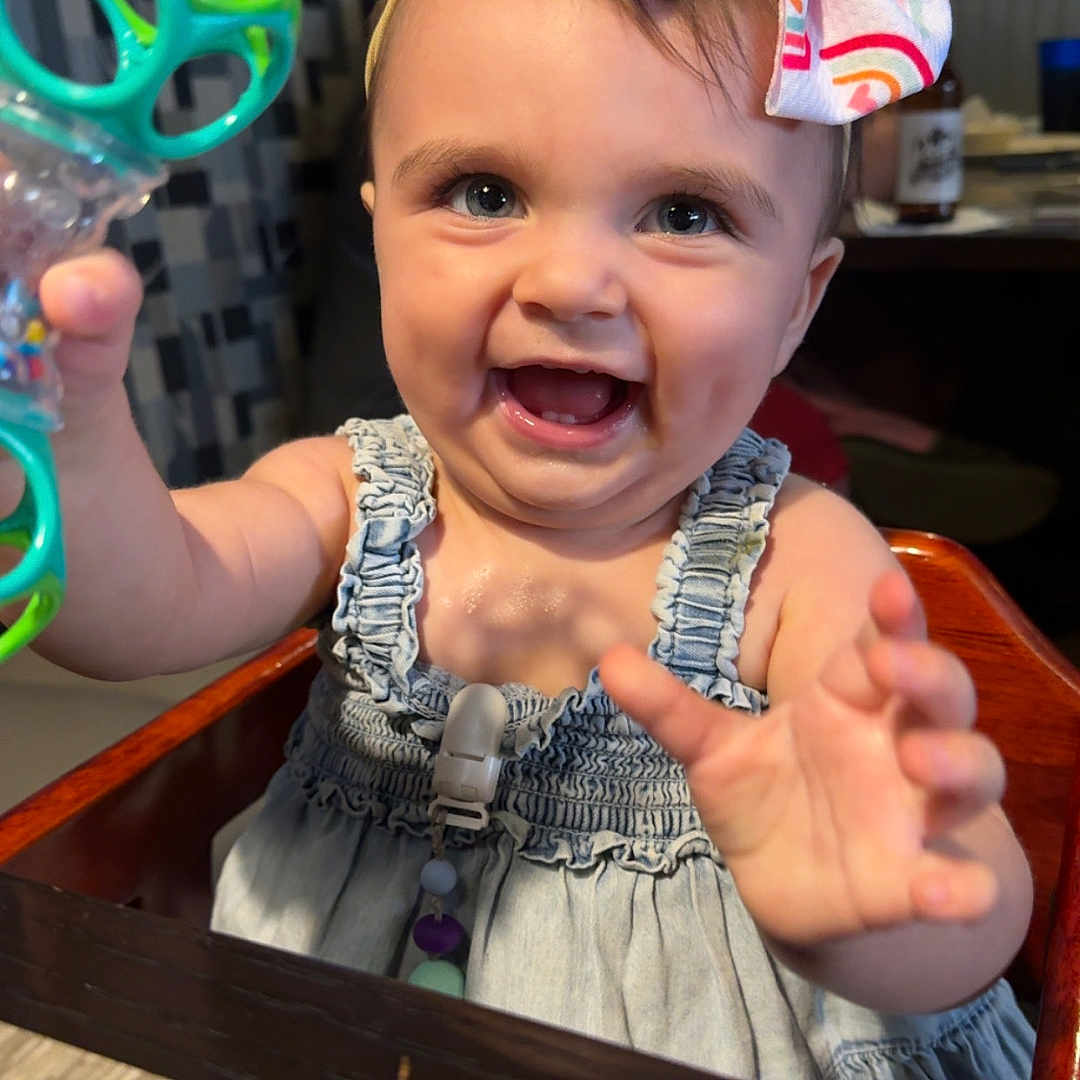Scarlett is registered to the contest to win money with this photo: baby, blue_eyes, bow, cheerful, child, clothing, cute, dress, face, furniture, hand, happy, headband, high_chair, indoor, person, skin, smiling, table, toy