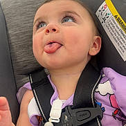 Scarlett is registered to the contest to win money with this photo: baby, child, tongue, car_seat, purple_clothing, straps, beads, indoor, store, blurred_background, cute, expression, person, seatbelt, face, head, infant, playful, smiling, closeup