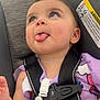 baby, child, tongue, car_seat, purple_clothing, straps, beads, indoor, store, blurred_background, cute, expression, person, seatbelt, face, head, infant, playful, smiling, closeup
