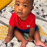 Dallas is registered to the contest to win money with this photo: baby, child, infant, red_shirt, blue_shorts, pacifier, bed, bedding, patterned_bedspread, curly_hair, sitting, indoors, looking_up, cute, young_child, soft_lighting, portrait, skin, expression, floor