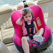 Freyja is registered to the contest to win money with this photo: beige, car_interior, car_seat, casual_clothing, child, curly_hair, daylight, footwear, person, pink, portrait, restraint, seat, seatbelt, sitting, sunlight, toddler, vehicle, window, young_child