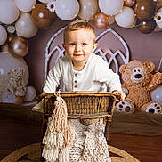 Léo a rejoint le concours — aidez-le/la à gagner de superbes lots ! toddler, child, wicker_chair, knit_blanket, balloons, white_balloons, bronze_balloons, plush_teddy_bear, wooden_floor, round_rug, smiling, indoor, decor, portrait, cute, baby, festive, happy, face, clothing