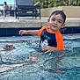 child, toddler, pool, water, swimming, splashing, orange_shirt, navy_shirt, smiling, face, wet_hair, outdoor, daylight, blurred_background, lounge_chair, plant, happy, person, recreation, summer