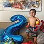 child, toddler, balloon, number_two, fish_balloon, striped_shorts, smiling, indoor, couch, pillow, abstract_painting, bare_chest, happy, home, playful, celebration, colorful, toy, living_room, decor
