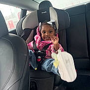 Dazyiere is registered to the contest to win money with this photo: automobile, backseat, black_hair, car_interior, car_seat, casual_clothing, child, cute, footwear, happy, jeans, person, pink_jacket, portrait, safety, seatbelt, sitting, smiling, white_bow, young_child