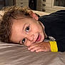 toddler, child, bed, smile, face, hand, curly_hair, black_clothing, yellow_cuff, indoor, portrait, resting, happy, person, closeup, young_child, bedroom, furniture, casual, cute