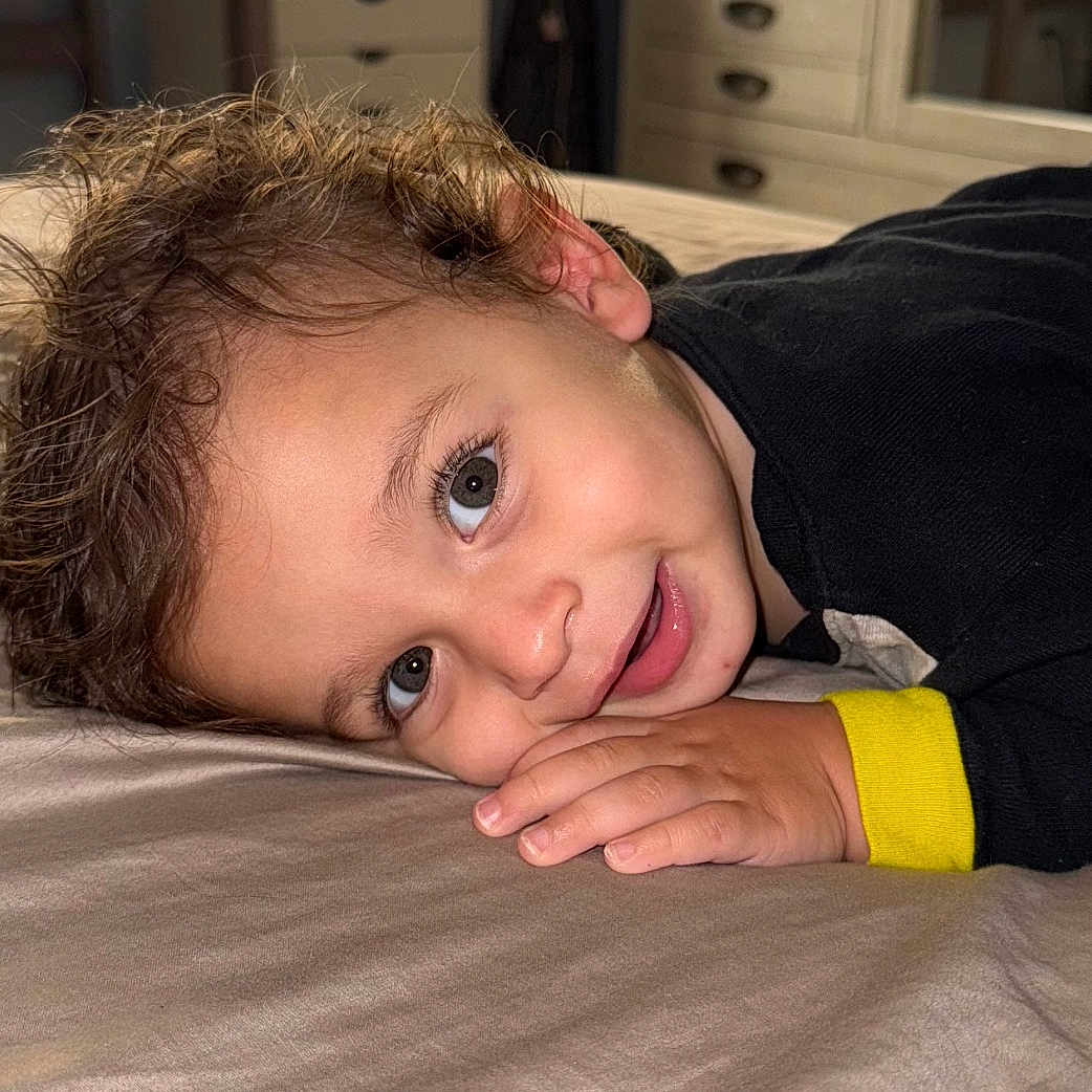 Sawyer joined the competition — help win amazing prizes! bed, bedroom, black_clothing, casual, child, closeup, curly_hair, cute, face, furniture, hand, happy, indoor, person, portrait, resting, smile, toddler, yellow_cuff, young_child