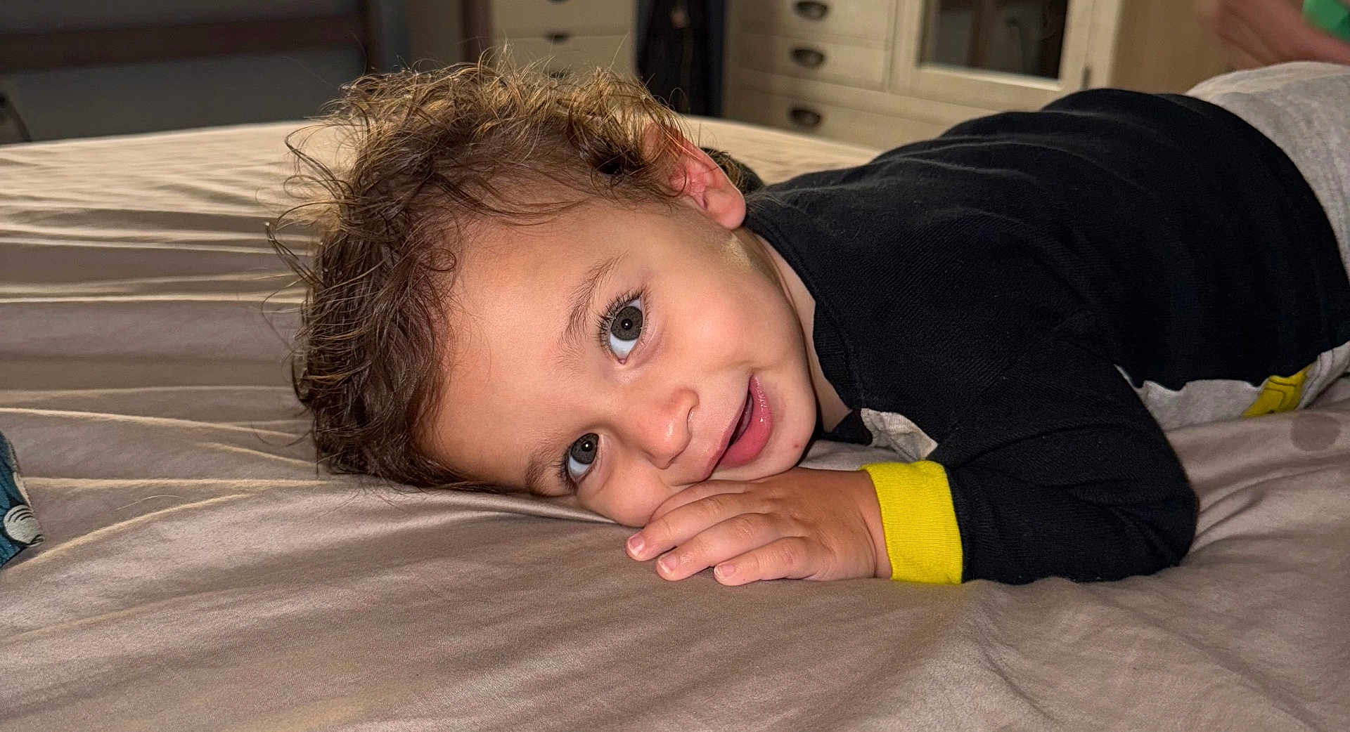 Sawyer joined the competition — help win amazing prizes! toddler, child, bed, smile, face, hand, curly_hair, black_clothing, yellow_cuff, indoor, portrait, resting, happy, person, closeup, young_child, bedroom, furniture, casual, cute