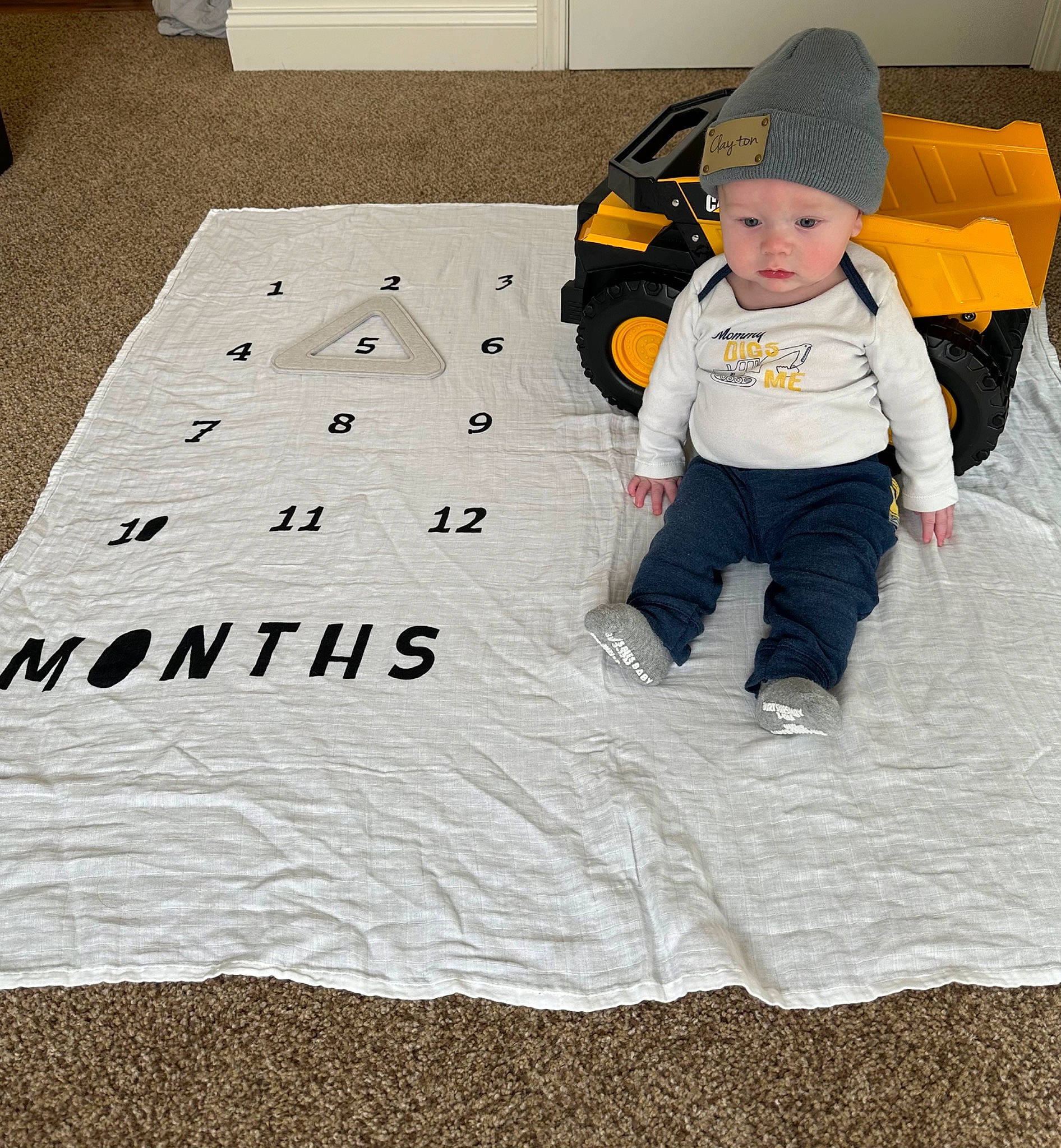 Clayton is registered to the contest to win money with this photo: baby, baby_toddler_clothing, beanie, cap, carmine, child, comfort, flooring, font, fun, headwear, hoodie, jacket, linens, pattern, person, personal_protective_equipment, sitting, sleeve, t_shirt