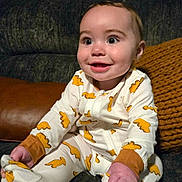Edith is registered to the contest to win money with this photo: baby, infant, smiling, onesie, dinosaur_print, couch, knitted_pillow, indoor, child, happy, cute, sitting, clothing, footwear, portrait, home, decor, furniture, face, person