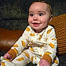 baby, infant, smiling, onesie, dinosaur_print, couch, knitted_pillow, indoor, child, happy, cute, sitting, clothing, footwear, portrait, home, decor, furniture, face, person