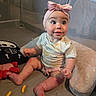 baby, child, infant, headband, pink_bow, sitting, snack, cheese_puffs, plush_toy, skeleton_toy, cushion, patterned_shirt, indoors, cute, happy, wide_eyes, curious, playpen, floor, person