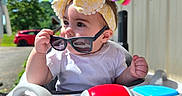 Edith is registered to the contest to win money with this photo: baby, car, child, colorful, daytime, expression, face, greenery, hand, headband, outdoor, play, seat, shirt, sunglasses, sunlight, toy, vehicle, walker, yellow