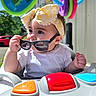 baby, child, sunglasses, headband, yellow, walker, toy, outdoor, sunlight, colorful, shirt, hand, face, expression, seat, vehicle, greenery, car, daytime, play