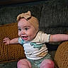 baby, child, smiling, headband, onesie, dinosaur_print, couch, pillows, knitted, yellow, gray, happy, sitting, indoor, person, cute, infant, arms_outstretched, feet, cozy