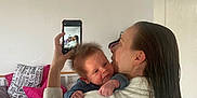 Henry is registered to the contest to win money with this photo: baby, woman, mirror, phone, selfie, bed, pillow, blanket, indoor, reflection, smile, hair, white_clothing, wooden_bedframe, text_on_pillow, hand, face, person, room, casual