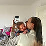 baby, woman, mirror, phone, selfie, bed, pillow, blanket, indoor, reflection, smile, hair, white_clothing, wooden_bedframe, text_on_pillow, hand, face, person, room, casual