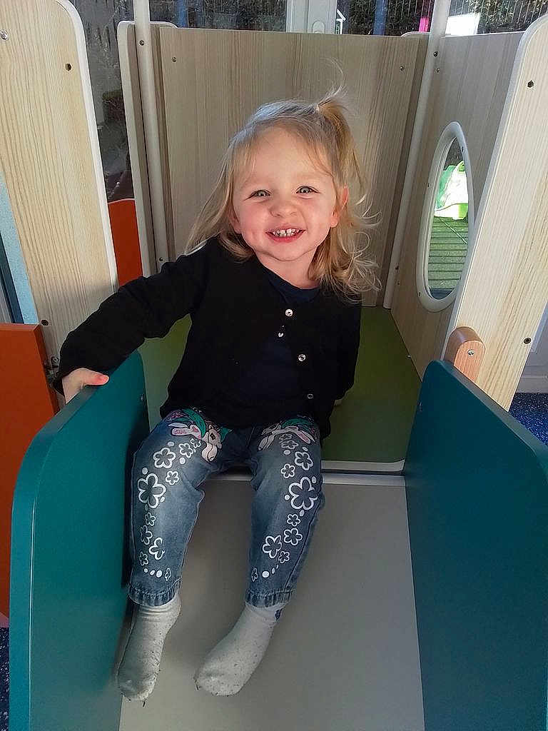 Catalyna a rejoint le concours — aidez-le/la à gagner de superbes lots ! blonde_hair, candid, cardigan, child, cute, happy, indoor_play, jeans, light, missing_tooth, play_equipment, playground, portrait, sitting, slide, smile, socks, toddler, window, wooden_panel