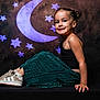 child, girl, smile, sitting, black_top, green_skirt, sneakers, stars, moon, background, portrait, indoor, studio, happy, face, hair, fashion, pose, dark_background, footwear
