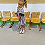 child, girl, glasses, braided_hair, striped_dress, sneakers, indoor, floor, chairs, wooden_chairs, playful_pose, smiling, young, person, cute, fashion, hair_tie, background, casual, happy