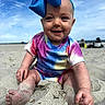 baby, child, sand, beach, smiling, colorful_clothing, bow, headband, outdoor, sunny, sky, people_in_background, happy, sitting, footwear_free, portrait, infant, summer, vacation, playful