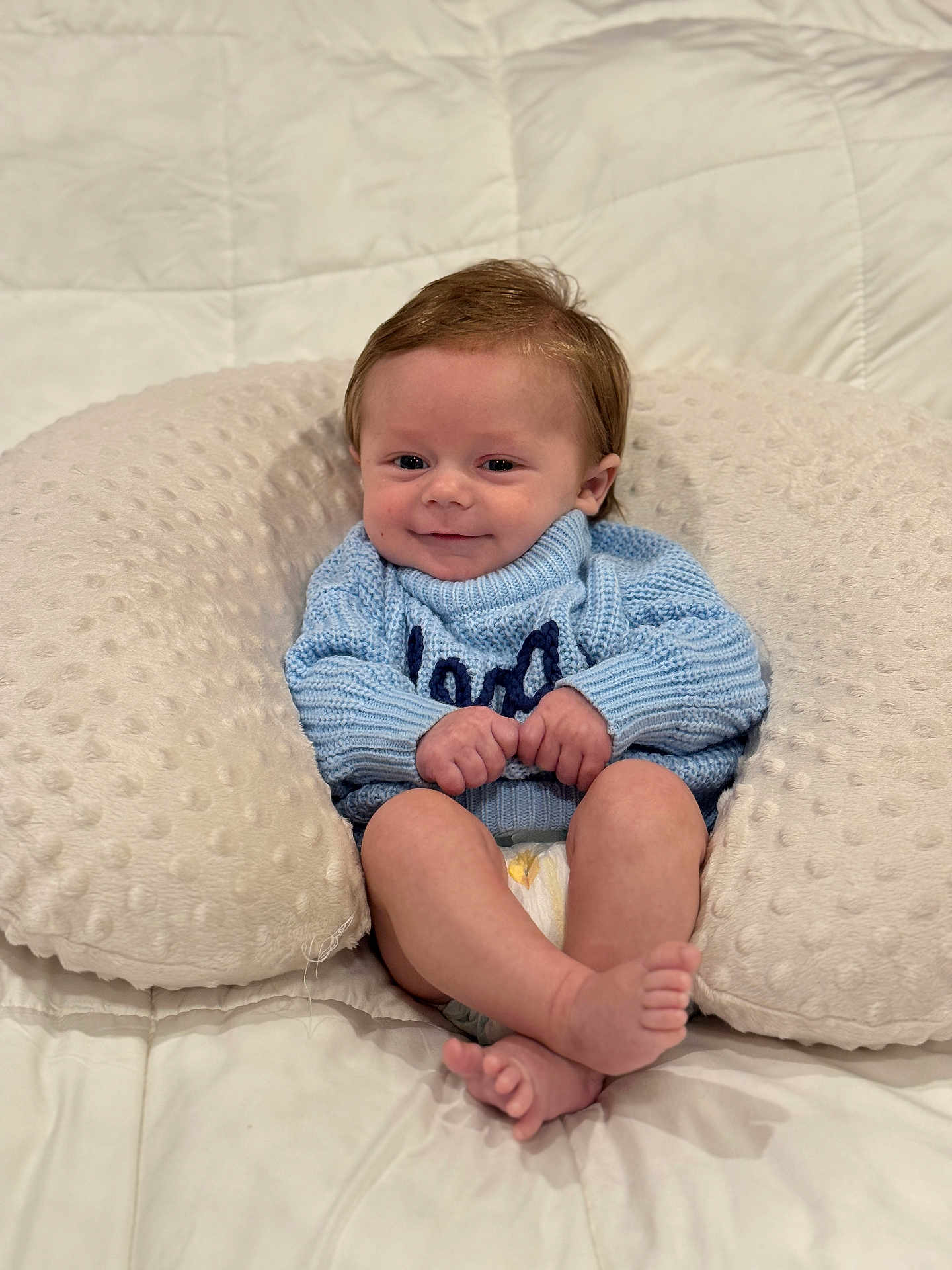 Logan joined the competition — help win amazing prizes! baby, infant, smile, blue_sweater, pillow, diaper, cute, cozy, soft_texture, bed, child, person, happy, indoors, skin, feet, hands, head, face, young