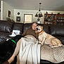 affection, black_dog, blanket, bookshelf, carpet, chandelier, cozy, cushion, dog, home_interior, lamp, living_room, pet, portrait, senior_person, side_table, slippers, sofa, tan_dog, woman