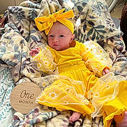 Willow is registered to the contest to win money with this photo: baby, infant, yellow_clothing, headband, bow, blanket, floral_pattern, plush, one_month, milestone, wooden_plaque, cute, newborn, portrait, indoors, soft_texture, baby_boy, baby_girl, clothing, baby_accessories