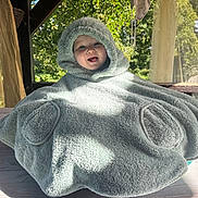 Charly a rejoint le concours — aidez-le/la à gagner de superbes lots ! baby, child, hooded_poncho, fluffy, smiling, outdoor, sunlight, wooden_table, greenery, happy, cute, cozy, portrait, infant, face, person, nature, daylight, warm_clothing, covered