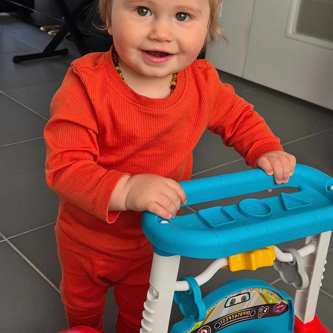 Jules participe au concours pour gagner de l'argent avec cette photo : toddler, child, smiling, orange_clothing, toy, walker, indoor, floor_tiles, baby, play, happy, young_child, person, furniture, colorful, plastic_toy, head, face, standing, cute
