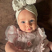 Lily joined the competition — help win amazing prizes! baby, infant, child, blue_eyes, headband, bow, pink_dress, frills, flower, beads, hand, soft_texture, brown_background, portrait, cute, young_child, laying_down, close_up, clothing, smile