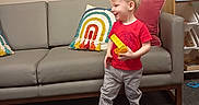 Ayden is registered to the contest to win money with this photo: child, boy, toy, red_shirt, gray_couch, rainbow_pillow, carpet, indoor, smiling, walking, painting, bird_art, furniture, home, living_room, sneakers, play, cute, happy, toddler