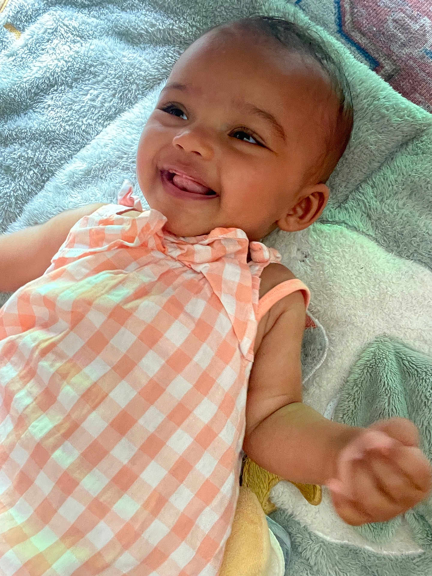 Safiya a rejoint le concours — aidez-le/la à gagner de superbes lots ! baby, child, smiling, happy, lying_down, dress, peach_color, checkered_pattern, blanket, soft_texture, infant, cute, face, skin, arm, person, indoors, closeup, portrait, playful