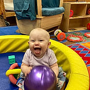 Giavanna is registered to the contest to win money with this photo: baby, child, infant, smiling, tongue_out, sitting, playroom, toy, ball, purple_ball, play_ring, colorful, indoor, daycare, wooden_furniture, chair, shelves, play_mat, happy, portrait