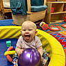 baby, child, infant, smiling, tongue_out, sitting, playroom, toy, ball, purple_ball, play_ring, colorful, indoor, daycare, wooden_furniture, chair, shelves, play_mat, happy, portrait