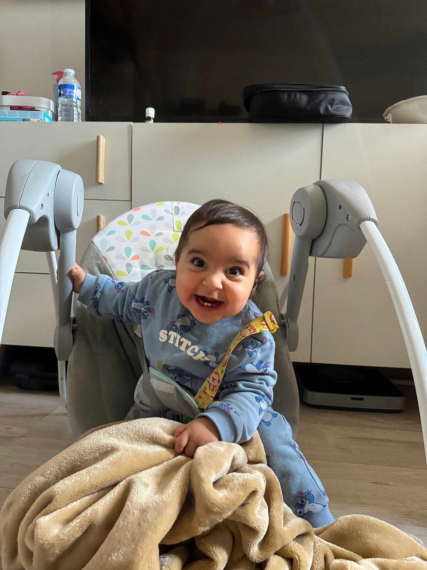 Aws participe au concours pour gagner de l'argent avec cette photo : baby, child, smiling, blue_clothing, blanket, baby_rocker, indoor, floor, cabinets, television, happy, person, toddler, toy_strap, cozy, cute, home, furniture, expression, casual