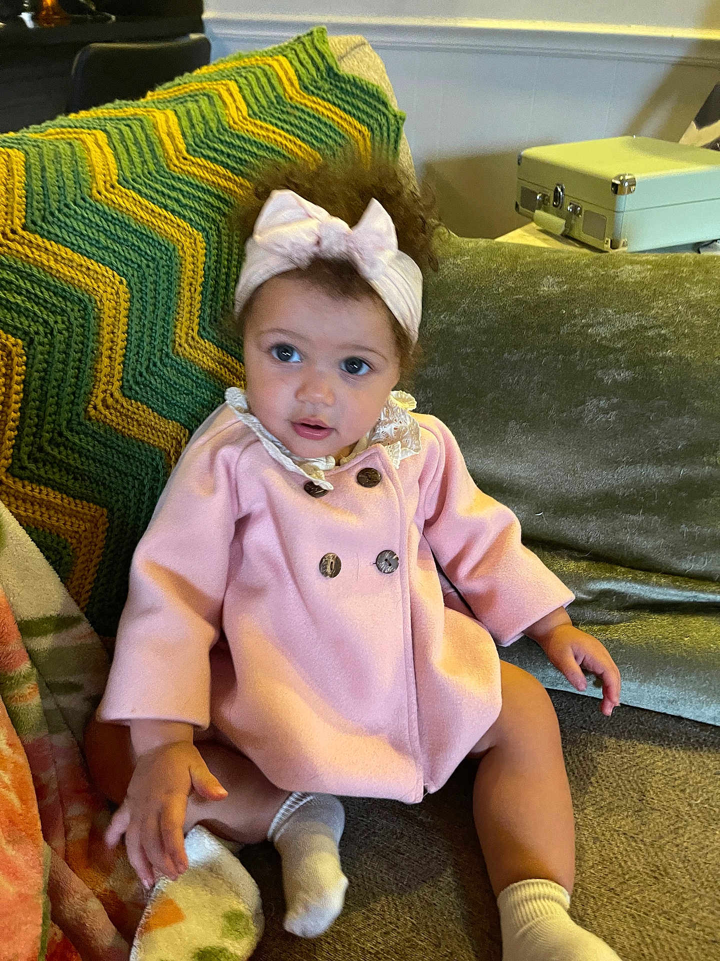 Penelope is registered to the contest to win money with this photo: baby, child, pink_coat, headband, couch, knitted_blanket, curly_hair, socks, indoors, seated, young_child, portrait, cute, looking_away, furniture, textile, cozy, home, person, infant