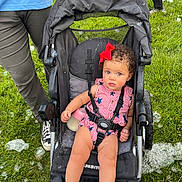 Penelope is registered to the contest to win money with this photo: toddler, child, stroller, baby_bottle, red_bow, curly_hair, blue_eyes, grass, bubbles, outdoor, person, white_shoes, socks, black_stroller, casual_clothing, summer, playful, family, fun, daytime