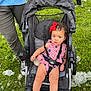 toddler, child, stroller, baby_bottle, red_bow, curly_hair, blue_eyes, grass, bubbles, outdoor, person, white_shoes, socks, black_stroller, casual_clothing, summer, playful, family, fun, daytime