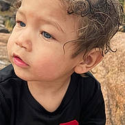 Keion joined the competition — help win amazing prizes! black_shirt, blue_eyes, casual_clothing, child, close_up, curly_hair, cute, face, greenery, natural_light, nature, outdoor, portrait, red_number, rocks, sitting, small_hands, thoughtful, toddler, young_child