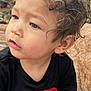 black_shirt, blue_eyes, casual_clothing, child, close_up, curly_hair, cute, face, greenery, natural_light, nature, outdoor, portrait, red_number, rocks, sitting, small_hands, thoughtful, toddler, young_child