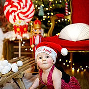 Léo a rejoint le concours — aidez-le/la à gagner de superbes lots ! baby, child, santa_hat, red_overalls, sled, christmas_tree, holiday_lights, wooden_floor, indoor, festive, decorations, toy, crawling, cute, face, person, christmas, red_chair, plush, winter